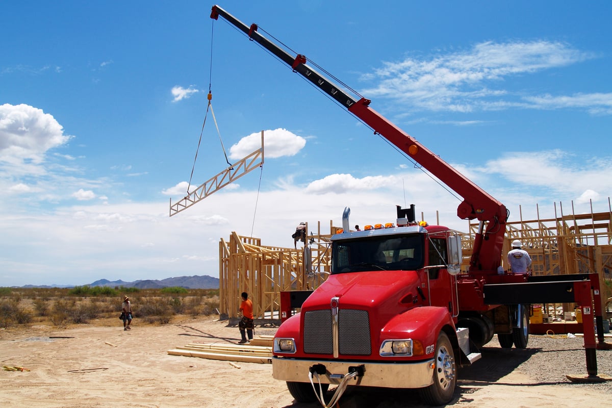 The image shows a crane truck hoisting a piece of framing into the air on a construction site.